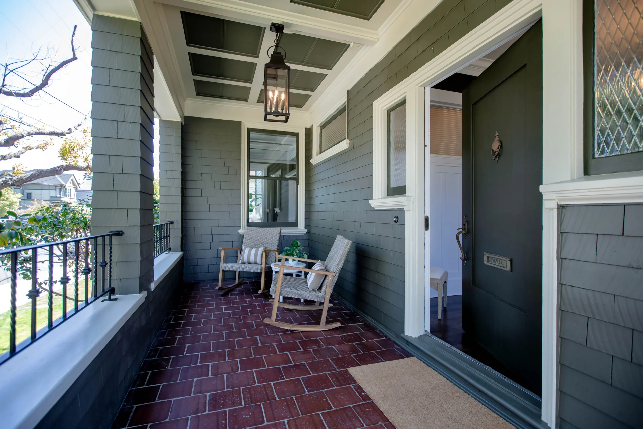Cozy front porch with chairs and hanging lantern, gray siding, and brick flooring.